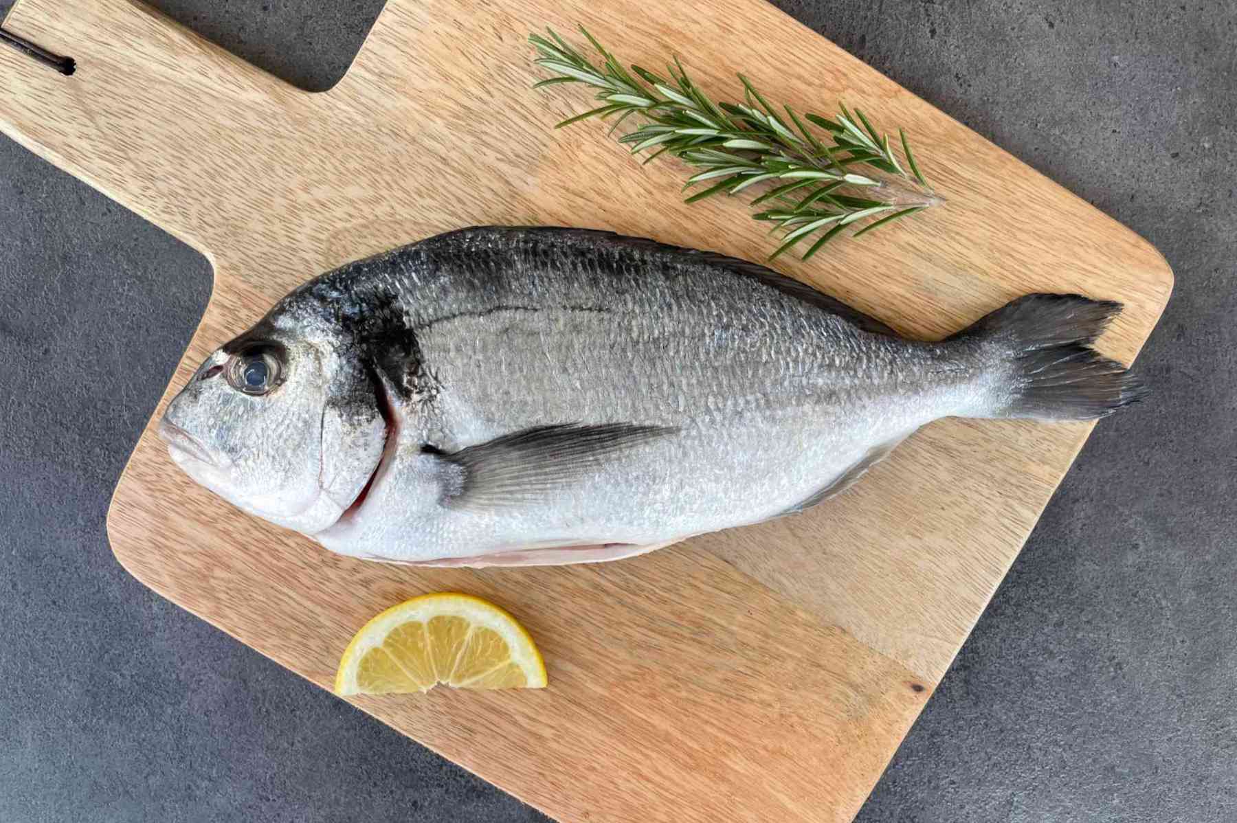 Frische ganze Dorade roh auf Holzbrett mit Rosmarin und Zitrone – ideal für mediterrane Fischgerichte